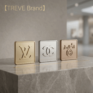 「【TREVE Brand】」のゴールドロゴが左上に配された、洗練されたモダンな空間。大理石のカウンターの上には、世界最高峰のブランドを象徴する3つのスクエア型メタルエンブレムが整然と並んでいる。左から「ルイ・ヴィトン（LV）」のゴールド、中央に「シャネル（ココマーク）」のシルバー、右に「エルメス（馬車ロゴ）」のブロンズが配され、各ブランドのアイデンティティを際立たせている。背景はミニマルで落ち着いたグレーのトーンで統一され、高松市のブランド買取専門店「TREVE」が、バッグや財布、アクセサリーの真贋から微細なコンディションまでを精密に鑑定し、国際相場に連動した最高値を提示するイメージ。