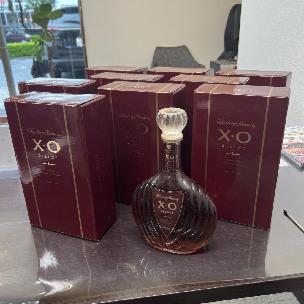 高松市のトレリバで大量買取したサントリーブランデーXOデラックス。ボルドー色の外箱の前に置かれた未開栓のシェル型ボトルとお酒の山。