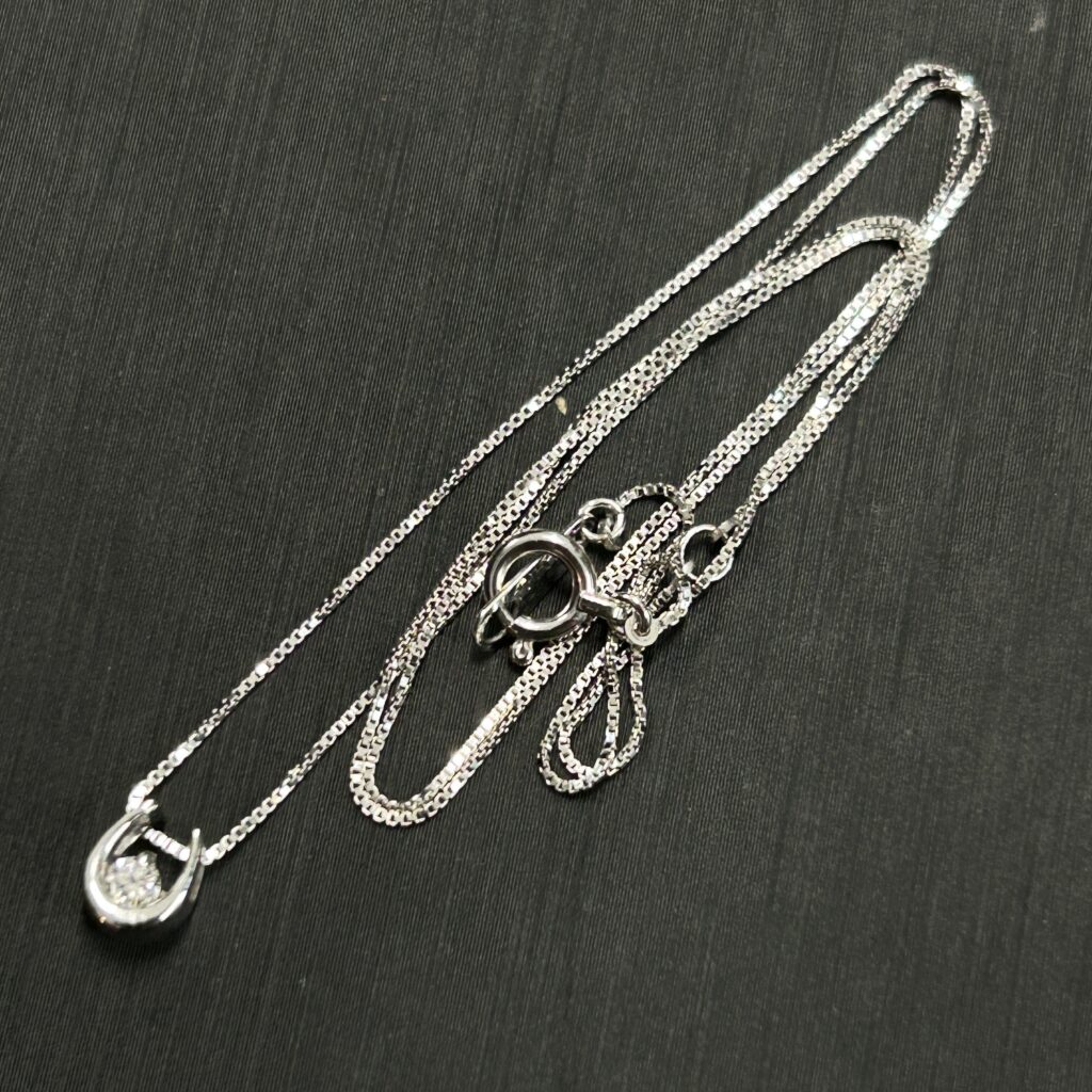 高松市のトレリバで買取したK10ホワイトゴールドのダイヤモンドネックレス。黒い背景の上に置かれた、ホースシュー（馬蹄）モチーフの一粒ダイヤジュエリーの画像。