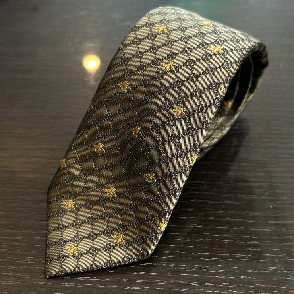高松市のトレリバで買取したグッチ（GUCCI）のシルクネクタイ。テーブルの上に丸めて置かれたモノグラムGGパターンとハチ（ビー）刺繍の画像。