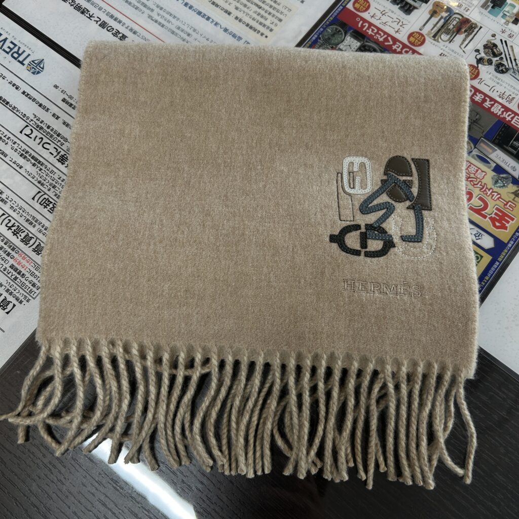 高松市のトレリバで買取したエルメス（HERMES）のカシミヤウールマフラー。テーブルの上に綺麗に畳んで置かれたベージュの本体とHパズル刺繍ロゴの画像。