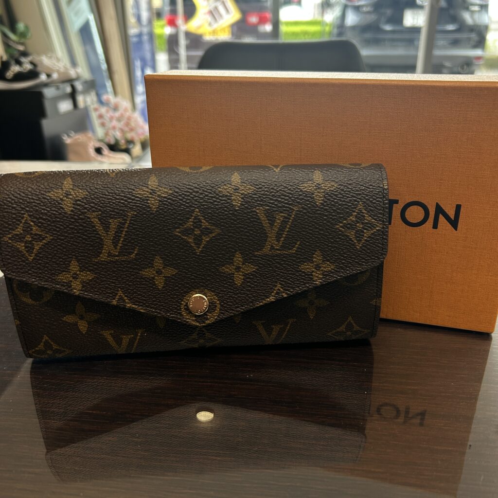 高松市のトレリバで買取したルイ・ヴィトン（LOUIS VUITTON）の長財布「ポルトフォイユ・サラ」。テーブルの上に置かれたモノグラムキャンバスの本体と、後ろにあるオレンジの純正箱の画像。