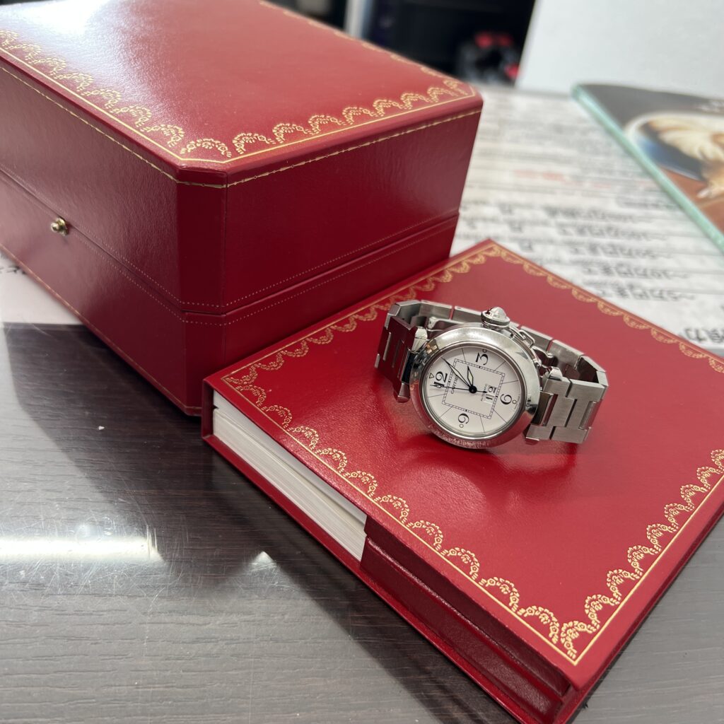 高松市のトレリバで買取したカルティエのパシャCビッグデイト。カルティエの赤いケースと冊子の上に置かれたステンレス自動巻き時計の画像。