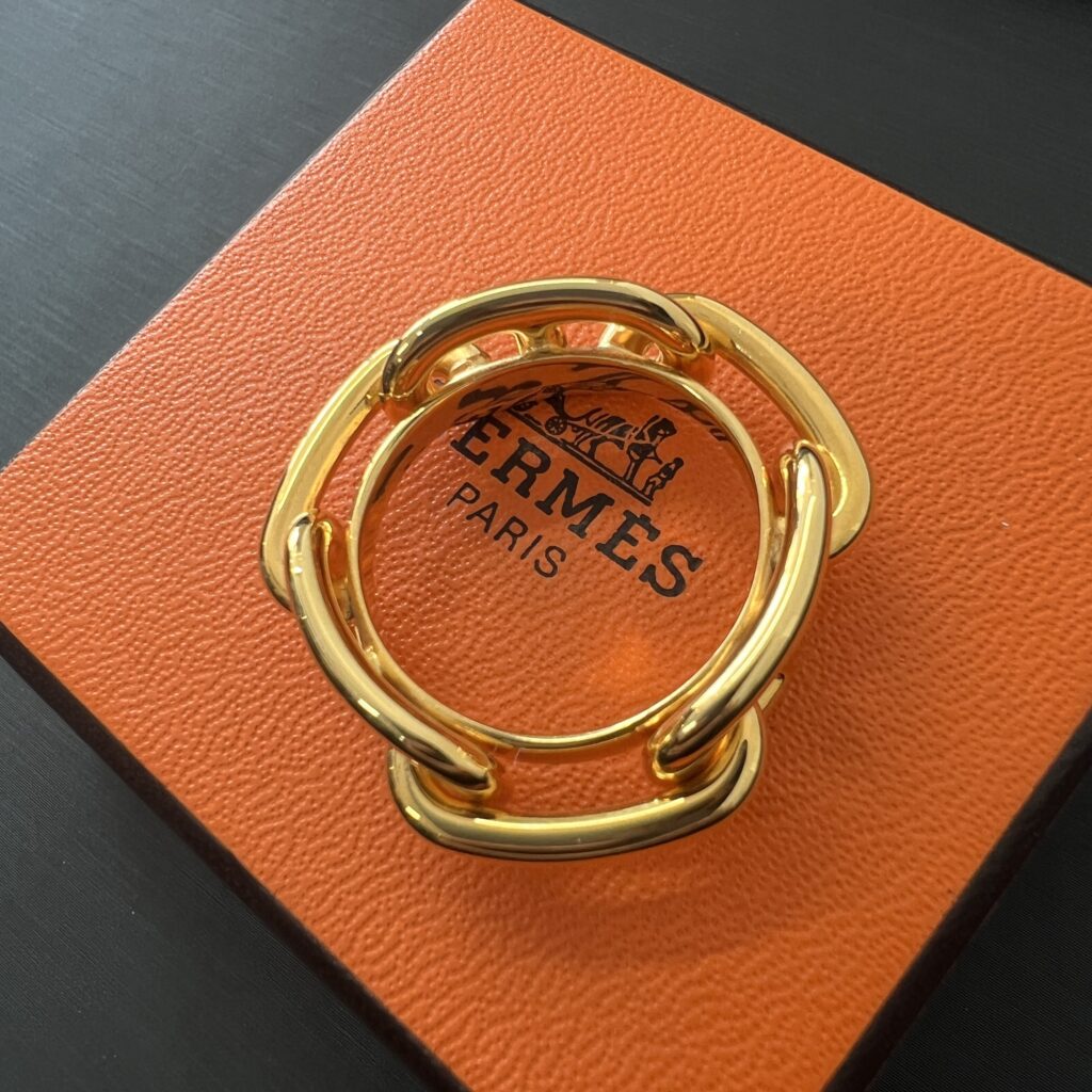 高松市のトレリバで買取したエルメスのシェーヌダンクルスカーフリング。オレンジのHERMES外箱の上に置かれたゴールドスカーフリングの画像。